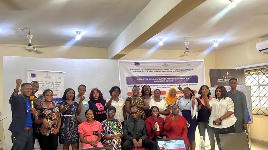 A group photo of facilitator and participants at a disability-inclusive SGBV response training workshop, standing and seated in a well-lit hall with banners behind them, smiling and raising their fists in solidarity, with presentation materials and a projector screen visible in front.