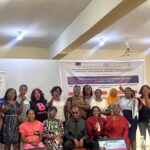 A group photo of facilitator and participants at a disability-inclusive SGBV response training workshop, standing and seated in a well-lit hall with banners behind them, smiling and raising their fists in solidarity, with presentation materials and a projector screen visible in front.