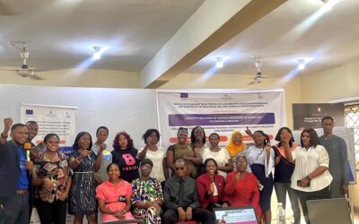 SARC Training Group Photo A group photo of facilitator and participants at a disability-inclusive SGBV response training workshop, standing and seated in a well-lit hall with banners behind them, smiling and raising their fists in solidarity, with presentation materials and a projector screen visible in front.
