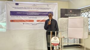 Melody Omosah Esq., Facilitator standing holding a mic, wearing a black long sleeve on an black trousers, with a pulpit having the NAPVID logo on it.