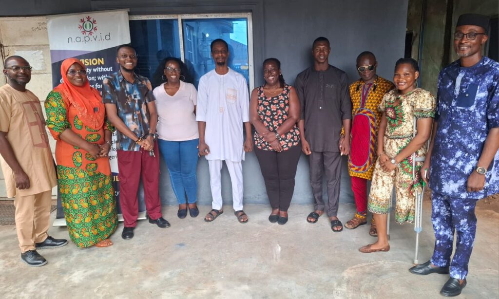 A group of nine people stand in a semi-circle outside a building beside a NAPVID banner. The banner displays information about disability rights, inclusion, and equal opportunities for persons with disabilities. The individuals are dressed in a mix of traditional and casual clothing, including patterned outfits, tunics, and a light-colored polo shirt. One person on the far right is using a forearm crutch. Behind the group is a gray wall with a large blue-tinted window. The photo appears to document a meeting, stakeholder engagement, or advocacy activity related to disability inclusion and community development.