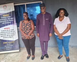 Three people stand side by side outside a building next to a vertical NAPVID banner. The banner displays the organization's vision, mission, and core values focused on promoting the rights, inclusion, and equal opportunities of persons with disabilities in Nigeria. One person in the center is dressed in a purple traditional outfit and dark glasses, while the other two wear casual clothing. A window and gray wall are visible in the background.